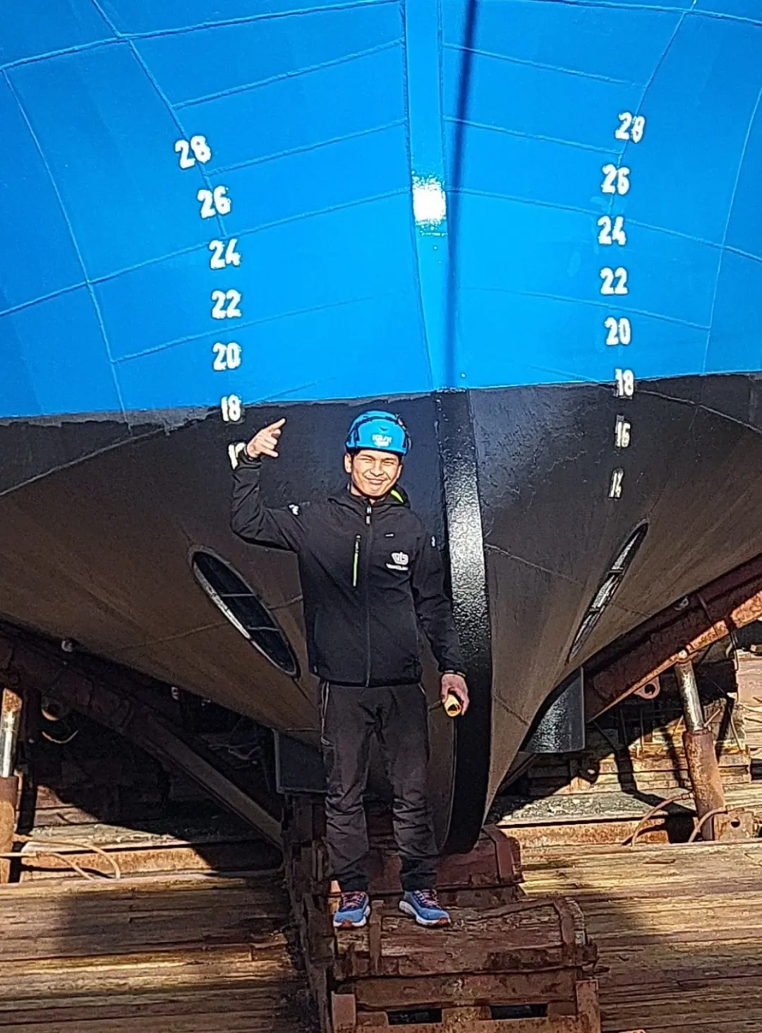 Smilande mannskap i Vestbjørn-jakke og hjelm under baugen på MS Vestbjørn, framfor djupgangsmerka på skroget